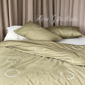 Può includere: Un set di biancheria da letto composto da un copripiumino e due federe in un colore verde oliva tenue, decorato con un motivo ripetitivo di cerchi bianchi disegnati a mano. Il set include una federa bianca tinta unita e il testo Alone Gifts Studio.