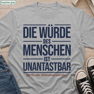 Op de afbeelding: Een heather grijs t-shirt met korte mouwen en donkerblauwe tekst "DIE WÜRDE DES MENSCHEN IST UNANTASTBAR" met de kleinere rode tekst "gilt für alle. ohne wenn und aber." onder de hoofdboodschap gedrukt.