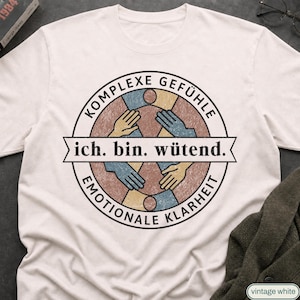 Puede incluir: Una camiseta de color crema con un gráfico circular de cuatro manos que se extienden hacia el centro. El texto dice KOMPLEXE GEFÜHLE, ich. bin. wütend. y EMOTIONALE KLARHEIT. El diseño utiliza tonos azules, beige y marrones apagados con textura.