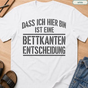 May include: A white short-sleeved t-shirt featuring dark grey distressed-style text that reads: DASS ICH HIER BIN IST EINE BETTKANTEN ENTSCHEIDUNG. The shirt has a crew neckline and a standard fit, with the text centred on the chest area.