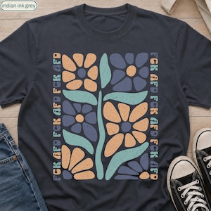Puede incluir: Una camiseta gris oscuro con un gráfico central de flores estilizadas en tonos naranja, azul y verde claro. El texto FCK AFD se repite verticalmente en una fuente desgastada a lo largo de los lados izquierdo y derecho del diseño floral.