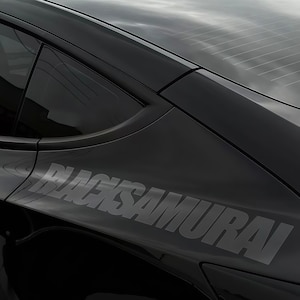Puede incluir: Primer plano del panel lateral de un coche negro con el texto "BLACKSAMURAI" en negrita gris. La ventana y el techo del coche son visibles, reflejando el cielo nublado.