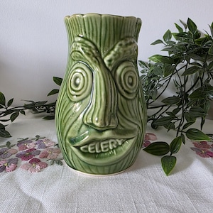 May include: A green ceramic vase shaped like a stalk of celery with a textured surface and a face featuring spiral eyes, a prominent nose, and a mouth with the word CELERY embossed on the lower lip. The glaze has a subtle crackle finish.