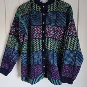May include: A long-sleeve knit cardigan featuring a multi-coloured patchwork pattern in shades of navy blue, forest green, purple, and teal. The garment has a round neckline, a front button closure with silver-toned buttons, and ribbed cuffs.