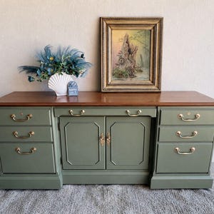 May include: A sage green wooden desk featuring a smooth brown top, six drawers, and a central cabinet with two doors. Each drawer and door is fitted with polished brass hardware. A framed landscape painting and a shell-shaped vase sit on top.