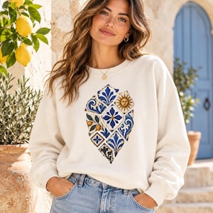 May include: A cream-colored crewneck sweatshirt featuring a central graphic print composed of various blue and white tile patterns, including floral motifs, a sun face, and citrus elements, arranged in a shield-like shape on the chest.