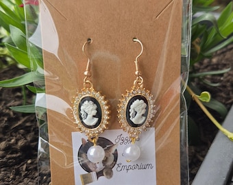 May include: A pair of gold-tone drop earrings featuring an oval black cameo with a white profile silhouette, surrounded by a border of small clear crystals. Each earring is finished with a single white pearl bead dangling from the bottom.