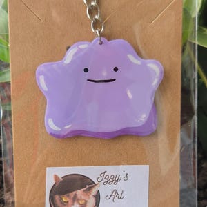 May include: A purple Ditto-shaped keychain with a silver chain. The keychain has a smiling face and is attached to a brown card with a sticker that reads "Izzy's Art Emporium". The background includes greenery and a red flower.