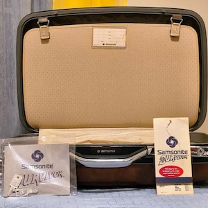 May include: A dark brown Samsonite Survivor hardshell carry-on suitcase with a tan interior lining. The open case features a central divider with an identification tag. Included are a plastic-wrapped manual and a vertical product information card.