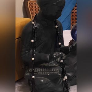 Pode incluir: Um colar longo com um fio de pequenas contas pretas facetadas intercaladas com pérolas brancas redondas. O colar está drapeado sobre uma escultura escura de acabamento fosco de uma figura sentada, exibindo um padrão de contas pretas e pérolas.