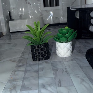 May include: Two small, artificial plants in decorative pots. One plant has long, green leaves and is in a black pot with a textured pattern. The other is a succulent in a white pot with a diamond pattern. The plants are on a marble-look surface.