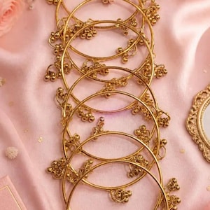 May include: Gold-colored bangles with floral accents are arranged on a pink satin surface. The bangles are adorned with small, round embellishments. The image has a soft, romantic aesthetic, with a book and flowers in the background.