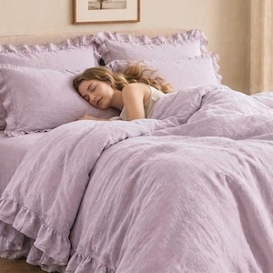 Puede incluir: Un juego de cama de color lavanda claro que incluye una funda nórdica y dos fundas de almohada a juego, todas adornadas con bordes decorativos de volantes. La tela tiene una apariencia suave y texturizada, creando un tono uniforme en todo el conjunto.