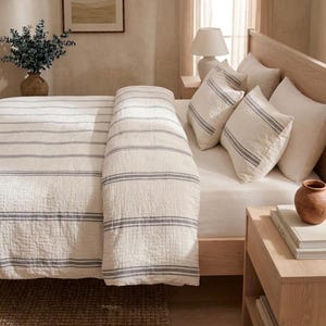 May include: A bed featuring a cream-coloured duvet cover and matching pillowcases, all decorated with thin, horizontal grey stripes. The bedding is arranged on a light-toned wooden bed frame, complemented by plain white pillows and a textured fabric finish.