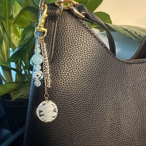 May include: A black textured handbag featuring a gold-toned metal clasp attached to a decorative charm. The charm consists of a gold chain with light blue beads, a clear glass bead with a white eye design, and a round clear glass pendant with white swirls.