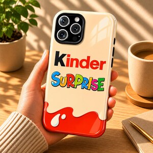Può includere: Custodia per telefono con la scritta "Kinder Surprise" in testo colorato. La custodia ha uno sfondo color crema con un design rosso e bianco nella parte inferiore. Le lenti della fotocamera del telefono sono visibili nella parte superiore.