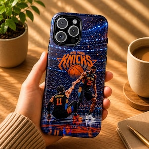 May include: A blue and orange New York Knicks phone case featuring a basketball court scene with two players. The case has the team's logo and player numbers. The case is held in a person's hand.