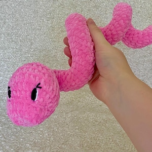 May include: A bright pink plush snake toy crafted from soft, textured yarn. The stuffed animal features a rounded head with black embroidered eyes and stitched eyelashes, and a long, winding body that curves in a serpentine shape.