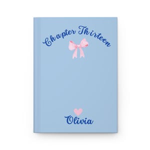 May include: A light blue hardcover journal featuring the dark blue text Chapter Thirteen arched over a pink ribbon bow illustration. Below the bow, a small pink heart sits above the dark blue name Olivia printed in a cursive script font.