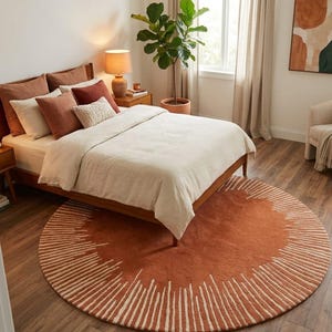 May include: A bedroom scene featuring a bed with a wooden frame, cream-colored bedding, and various brown and cream pillows. A round, rust-colored rug with white linear accents sits on the floor. A potted plant and a lamp are also visible.