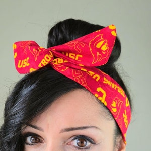 May include: A woman with dark curly hair wearing a red headband with the words "USC Trojans" and a yellow flame design.