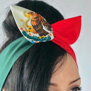 May include: A red, white, and green headband with a bow. The headband features the Mexican coat of arms on a white background. The design is a patriotic accessory, perfect for celebrating Mexican heritage or cultural events.