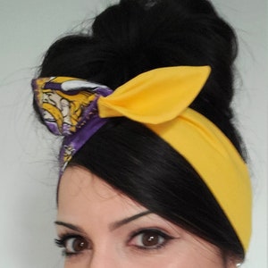 May include: A woman with dark hair styled in a bun wears a yellow and purple headband with a Viking logo. She is wearing a striped shirt and has a neutral expression.