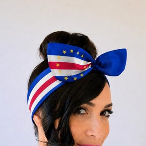 May include: A fabric headband featuring a bow detail with the Cape Verde flag design, including a blue background, a horizontal red stripe bordered by white, and a circular arrangement of ten yellow stars on the blue section of the bow.