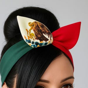 Puede incluir: Una diadema con una banda verde en el lado izquierdo y una banda roja en el derecho. En el centro, un lazo grande muestra una secci&oacute;n color crema con una ilustraci&oacute;n detallada de un &aacute;guila sobre un cactus, junto a una secci&oacute;n roja s&oacute;lida.