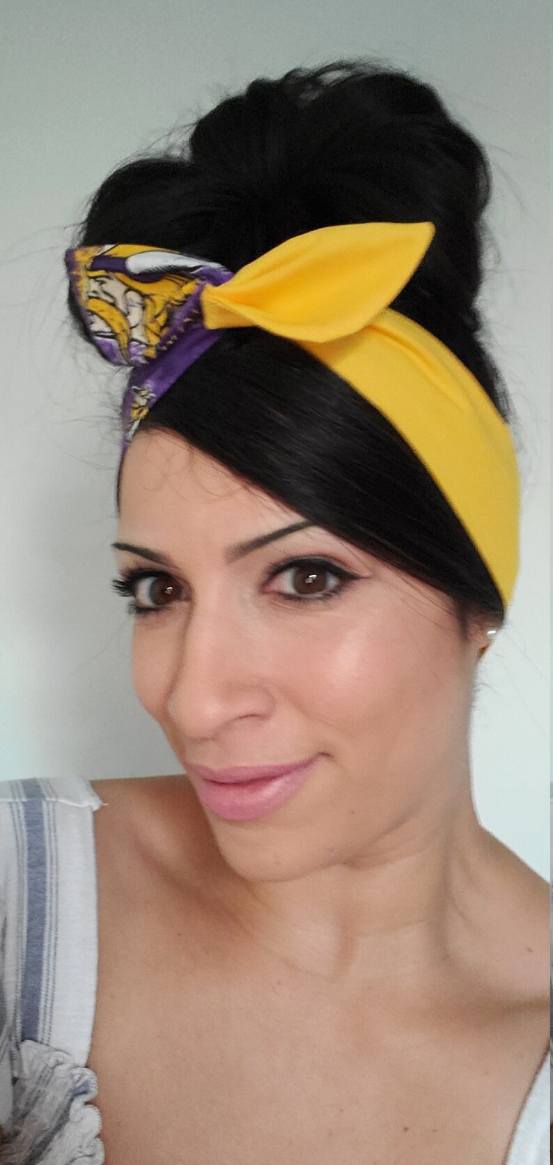 May include: A woman with dark hair styled in a bun wears a yellow and purple headband with a Viking logo. She is wearing a striped shirt and has a neutral expression.