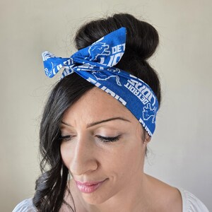 May include: A blue and white headband with the Detroit Lions logo and the text "Detroit Lions" repeated on the headband. The headband is tied in a bow on top of a person's head.