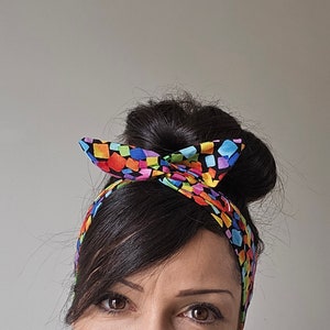 Op de afbeelding: Een kleurrijk hoofdband met een geometrisch patroon van vierkanten in verschillende kleuren, waaronder roze, blauw, groen, geel en oranje, op een zwarte achtergrond. Het hoofdband is bovenop het hoofd in een strik geknoopt.