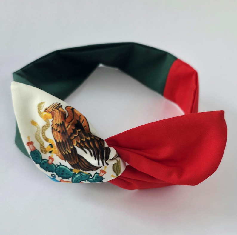 Puede incluir: Una diadema de tela anudada con un dise&ntilde;o tricolor en verde, blanco y rojo. La secci&oacute;n blanca muestra una ilustraci&oacute;n detallada de un &aacute;guila marr&oacute;n posada sobre un cactus verde, similar al emblema central de la bandera nacional mexicana.
