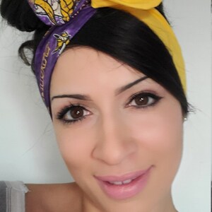 May include: A woman with dark hair tied up in a bun wears a purple and yellow headband with a team logo. The headband is tied in a bow at the top of her head. She is smiling and looking at the camera.
