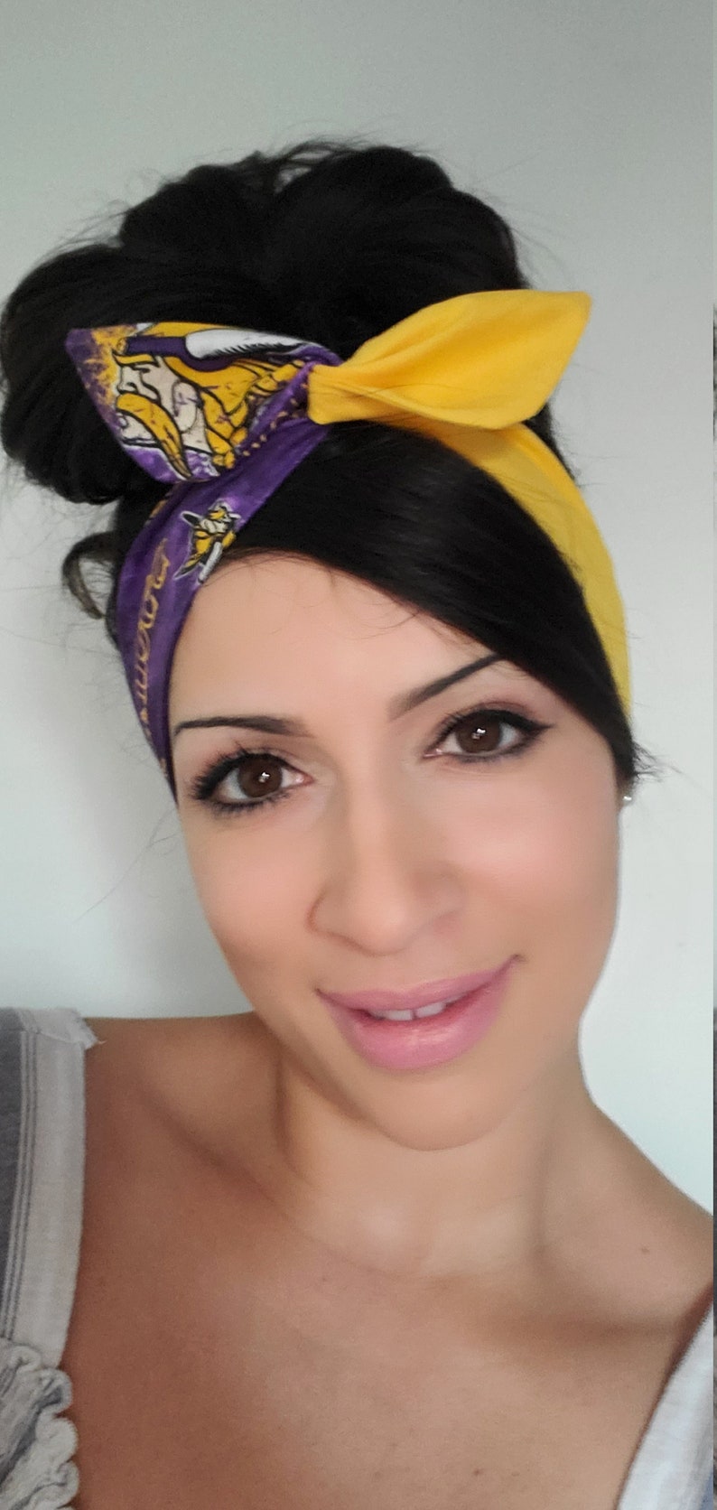 May include: A woman with dark hair tied up in a bun wears a purple and yellow headband with a team logo. The headband is tied in a bow at the top of her head. She is smiling and looking at the camera.