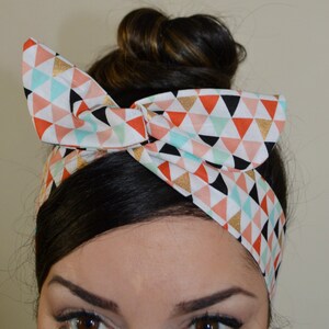Pode incluir: Uma faixa de cabelo colorida com laço, com um padrão geométrico de triângulos em coral, menta, preto e dourado sobre um fundo branco. A faixa de cabelo é usada na cabeça.