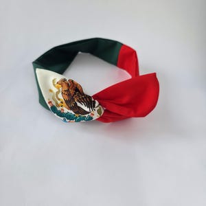 Puede incluir: Una diadema de tela con dise&ntilde;o anudado y un patr&oacute;n de bloques de color verde oscuro, blanco y rojo. La secci&oacute;n blanca muestra un estampado gr&aacute;fico detallado de un &aacute;guila posada sobre un cactus, replicando el emblema central de la bandera mexicana.