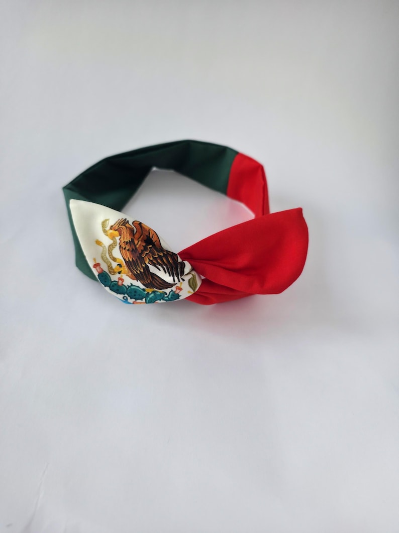 Puede incluir: Una diadema de tela con dise&ntilde;o anudado y un patr&oacute;n de bloques de color verde oscuro, blanco y rojo. La secci&oacute;n blanca muestra un estampado gr&aacute;fico detallado de un &aacute;guila posada sobre un cactus, replicando el emblema central de la bandera mexicana.
