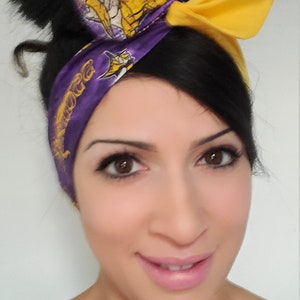 May include: A woman with dark hair wearing a purple and yellow headband with a team logo and the word "Vikings" on it. She is smiling and looking at the camera.