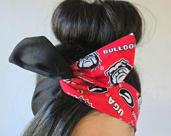 May include: A red and black headband with a bow. The red fabric features a repeating pattern of the University of Georgia Bulldogs logo and the text "UGA" and "BULLDOGS". The bow is black.