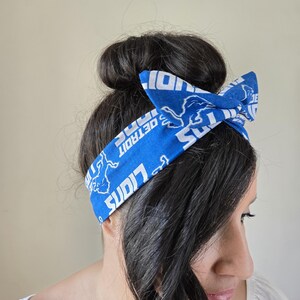 May include: A blue and white headband with the Detroit Lions logo and the words "Detroit Lions" repeated. The headband is tied in a bow on top of the head.