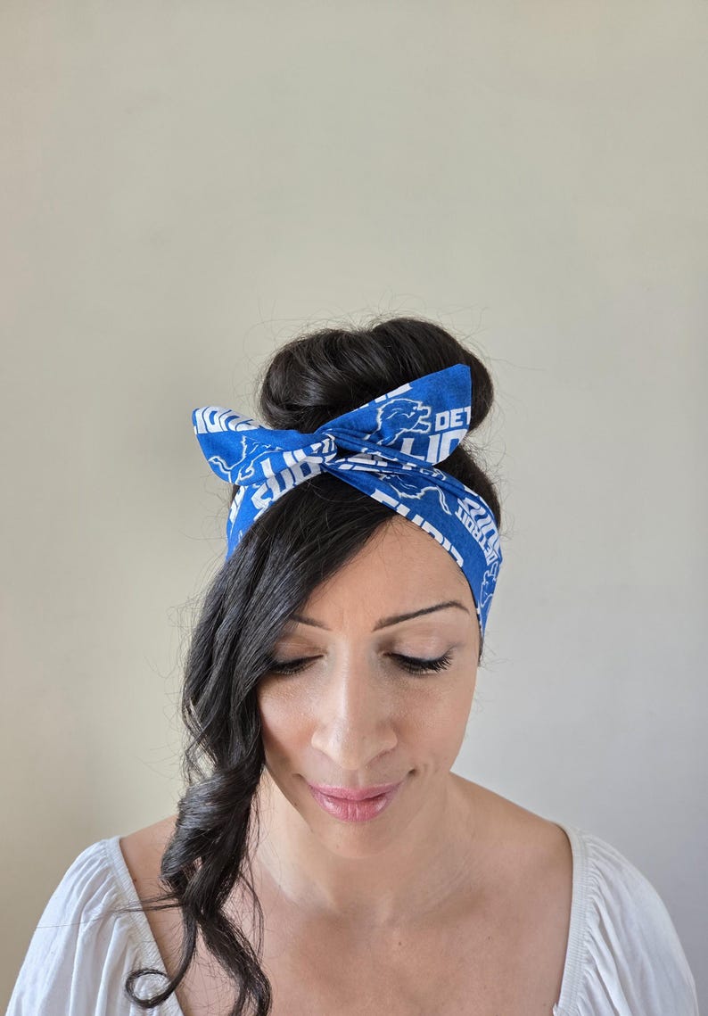 May include: A blue headband with a white print of the Detroit Lions logo and the words "Detroit Lions". The headband is tied in a bow on top of a woman's head.