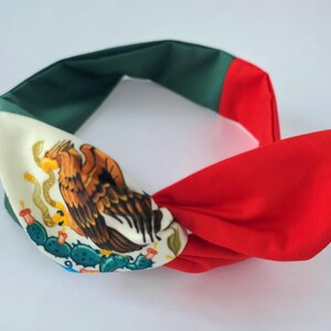 Puede incluir: Una diadema de tela con un dise&ntilde;o de bloques de color en verde, blanco y rojo. La secci&oacute;n blanca muestra un gr&aacute;fico detallado de un &aacute;guila posada sobre un cactus, sosteniendo una serpiente en su pico, similar al emblema de la bandera mexicana.