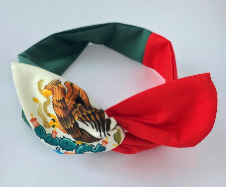 Puede incluir: Una diadema de tela con un dise&ntilde;o de bloques de color en verde, blanco y rojo. La secci&oacute;n blanca muestra un gr&aacute;fico detallado de un &aacute;guila posada sobre un cactus, sosteniendo una serpiente en su pico, similar al emblema de la bandera mexicana.