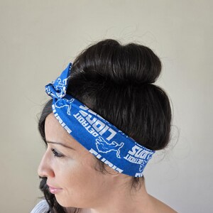 May include: A blue headband with a white print of the Detroit Lions logo and the words "Detroit Lions" repeated. The headband is tied in a knot on top of the head.