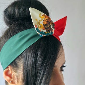 Puede incluir: Una diadema de tela con una banda ancha de color verde oscuro, una secci&oacute;n lateral de color rojo brillante y un detalle de nudo central en color blanco roto. La secci&oacute;n blanca muestra un gr&aacute;fico detallado de un &aacute;guila sobre un cactus.