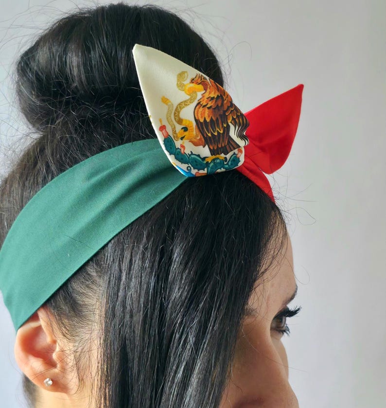 Puede incluir: Una diadema de tela con una banda ancha de color verde oscuro, una secci&oacute;n lateral de color rojo brillante y un detalle de nudo central en color blanco roto. La secci&oacute;n blanca muestra un gr&aacute;fico detallado de un &aacute;guila sobre un cactus.