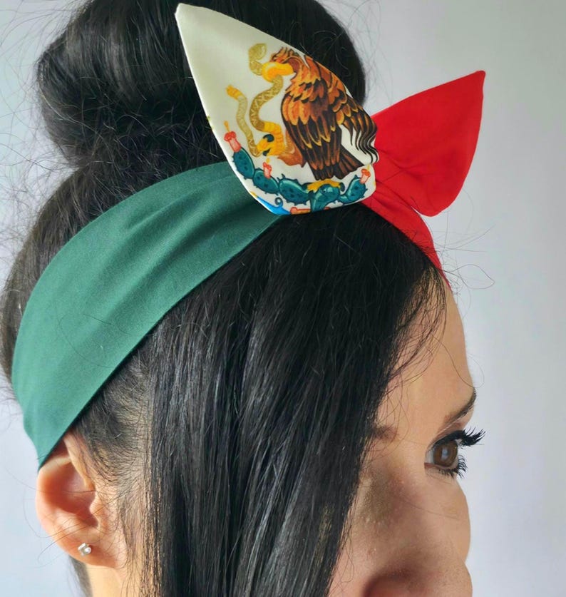 Puede incluir: Una diadema de tela con una secci&oacute;n verde oscuro s&oacute;lida y una secci&oacute;n roja, acentuada con un detalle de nudo de tela blanca que muestra una ilustraci&oacute;n gr&aacute;fica de un &aacute;guila marr&oacute;n posada sobre un cactus mientras sostiene una serpiente.