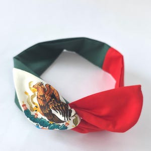 Puede incluir: Una diadema de tela con un dise&ntilde;o de bloques de color en verde oscuro, blanco y rojo. La secci&oacute;n blanca muestra una ilustraci&oacute;n detallada de un &aacute;guila posada sobre un cactus, replicando el emblema central de la bandera nacional mexicana.