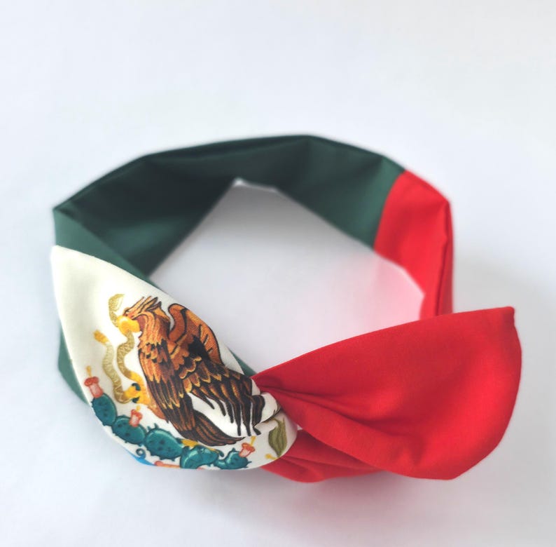 Puede incluir: Una diadema de tela con un dise&ntilde;o de bloques de color en verde oscuro, blanco y rojo. La secci&oacute;n blanca muestra una ilustraci&oacute;n detallada de un &aacute;guila posada sobre un cactus, replicando el emblema central de la bandera nacional mexicana.